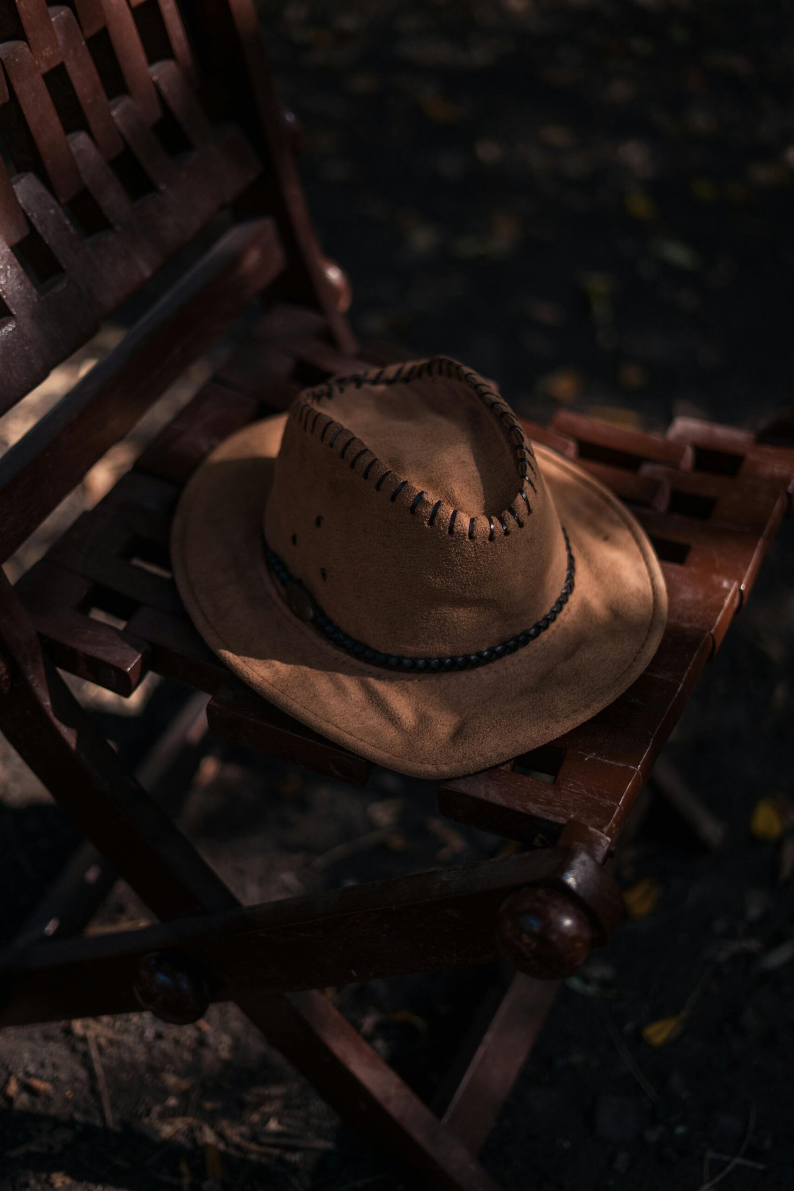 A cowboy hat rests on a wooden chair, casting shadows in outdoor light. Rustic and vintage feel.