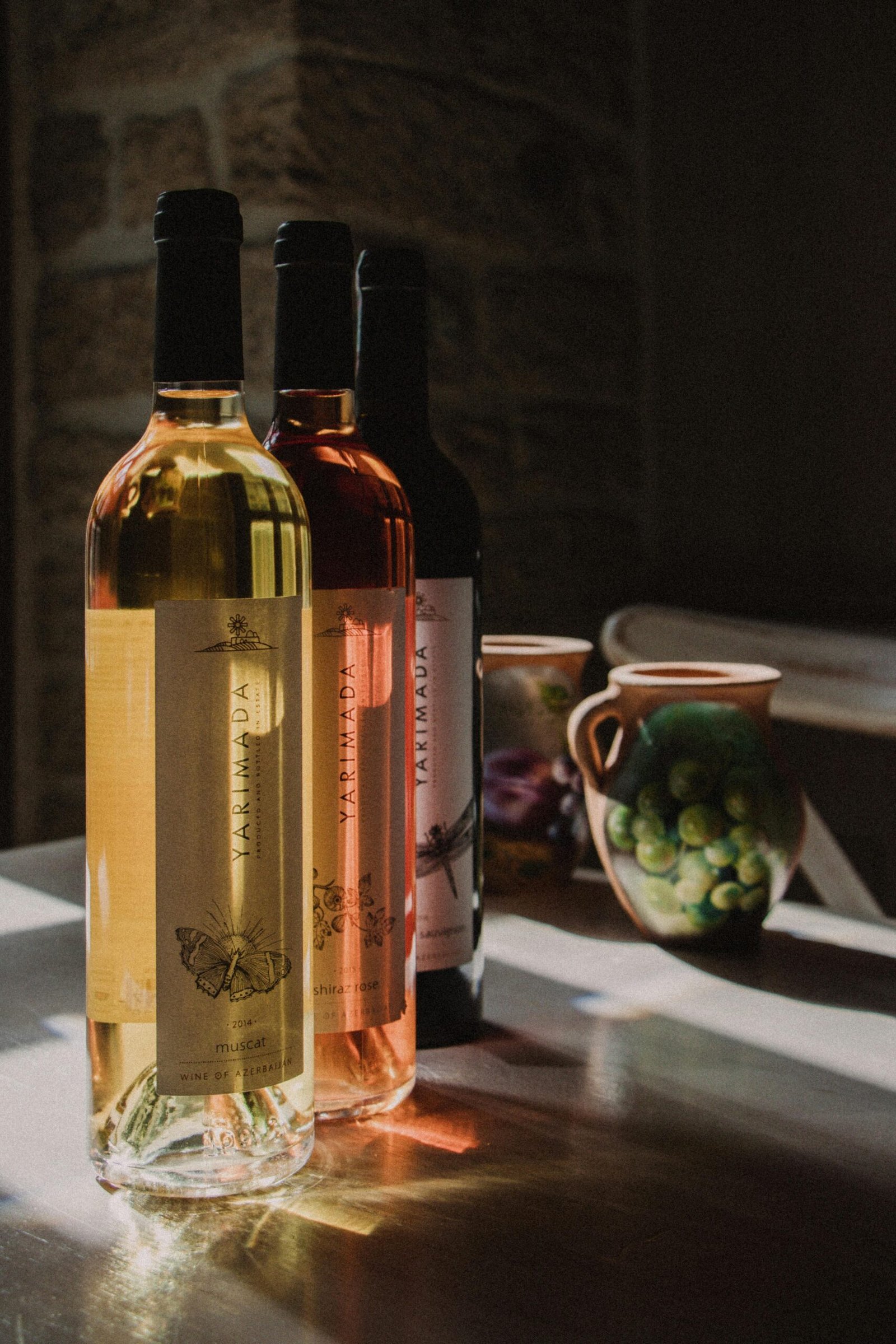 Three wine bottles on rustic table in sunlight, featuring white, red, and rosé wines.