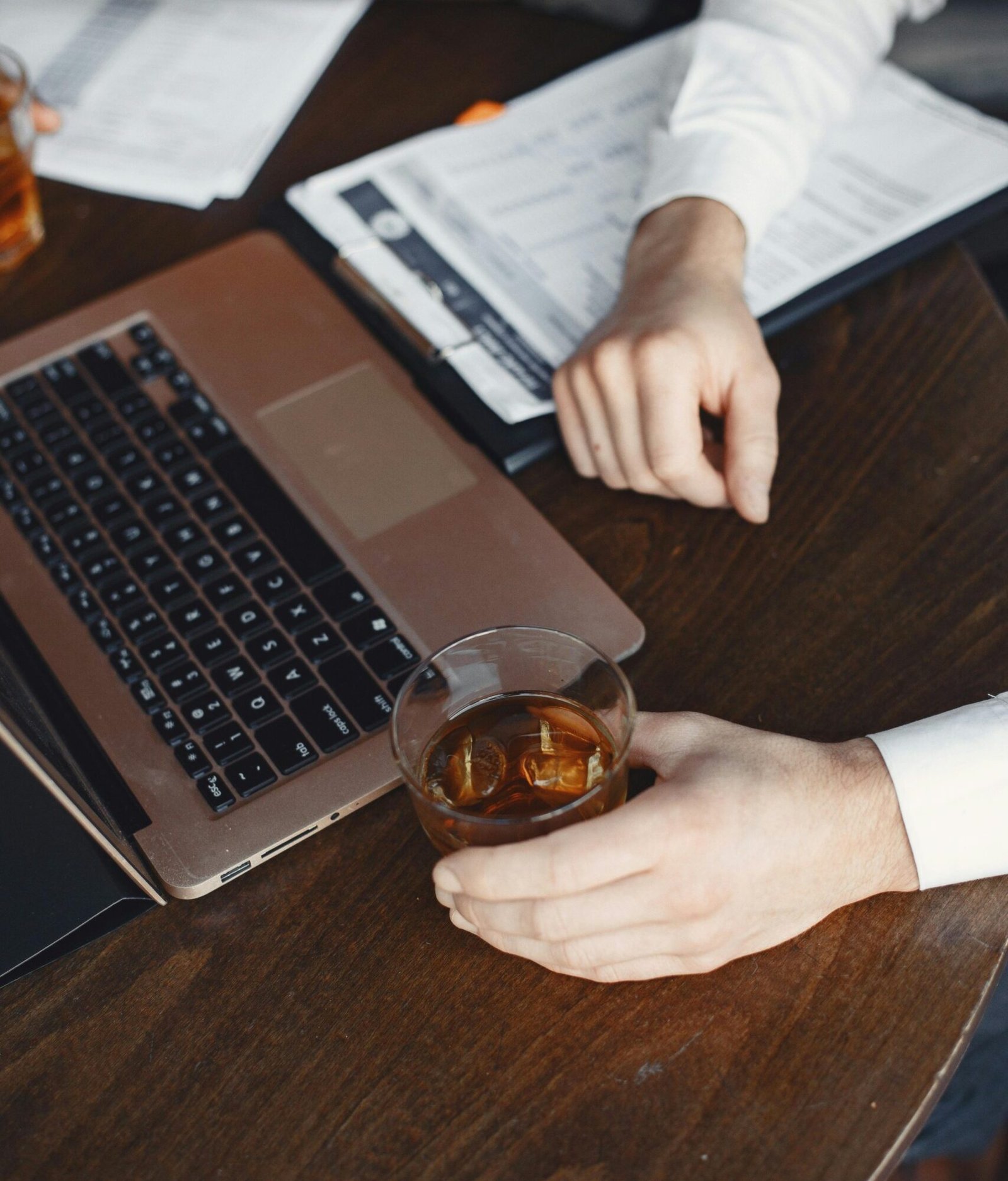Close-up of a businessman at a desk with a laptop, documents, and a glass of whiskey, conveying work and leisure.